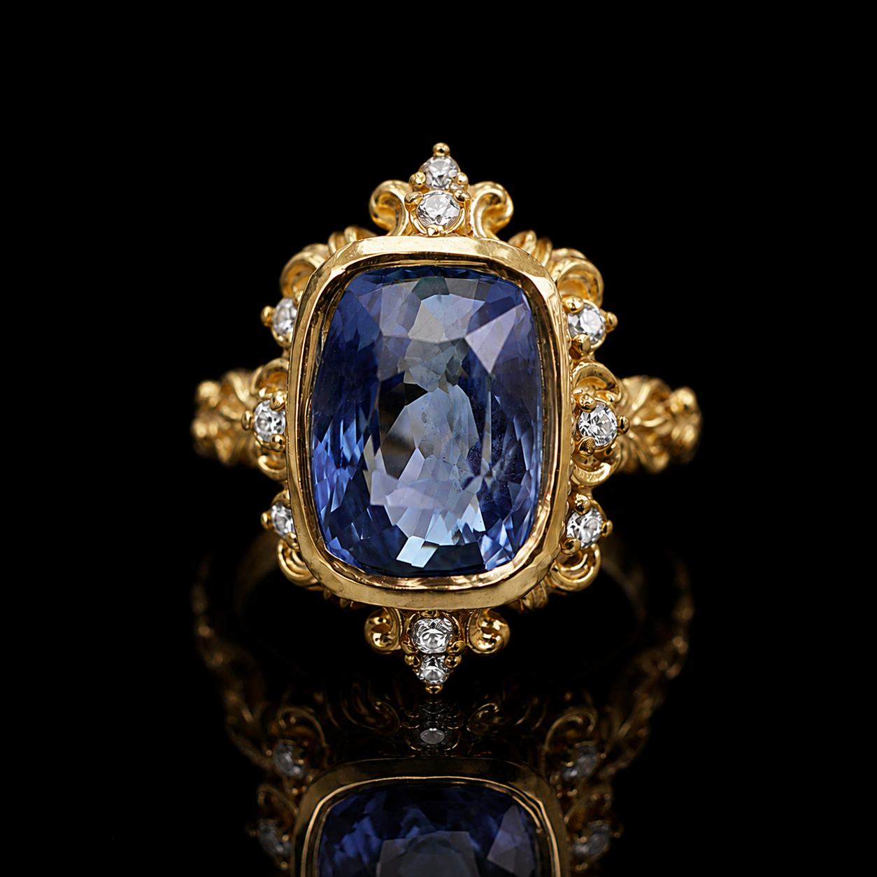 An ornate yellow gold ring featuring a large cushion-cut blue sapphire center stone, surrounded by small round diamonds and decorative scrollwork, set against a solid black background with a reflective surface below.