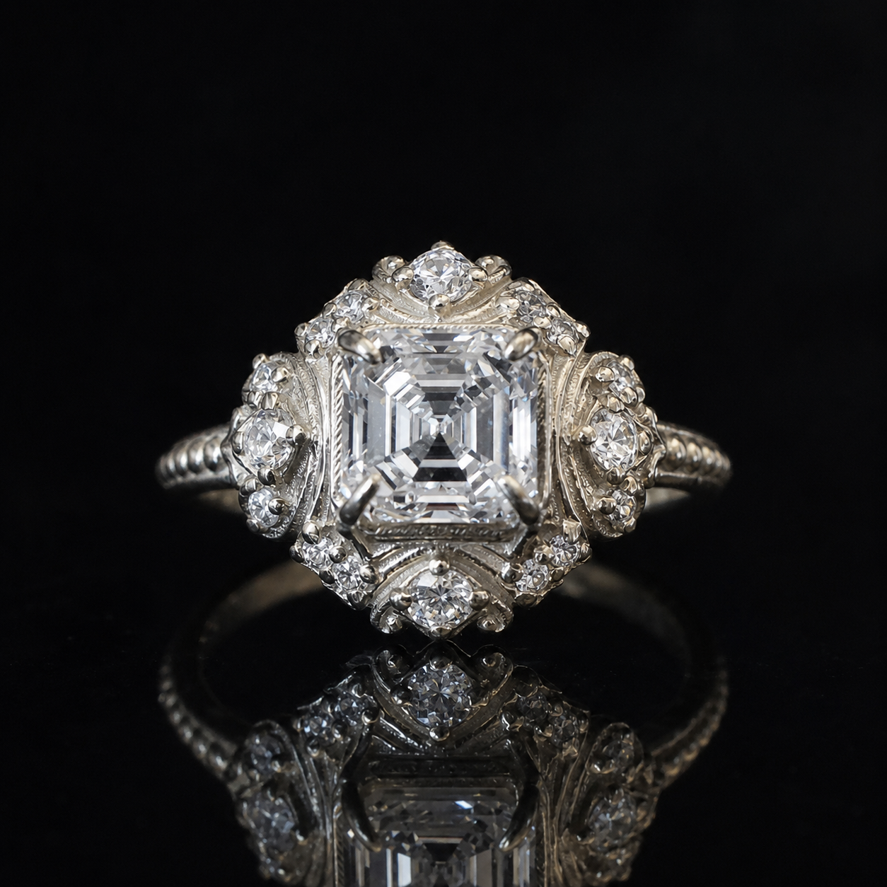 An ornate white gold diamond ring featuring a large emerald-cut center diamond surrounded by smaller diamonds in a decorative floral cluster setting, displayed on a reflective black surface.