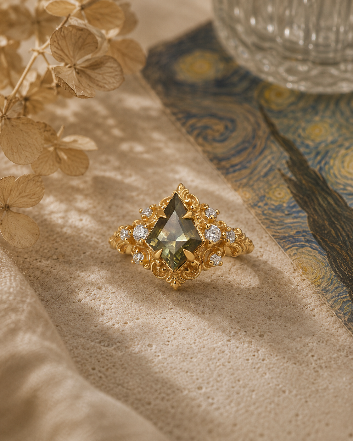 A gold ring with a kite shape bicolor sapphire center stone and small clear accent stones, styled on a textured beige fabric with dried flowers and a blue-and-gold patterned cloth in the background.