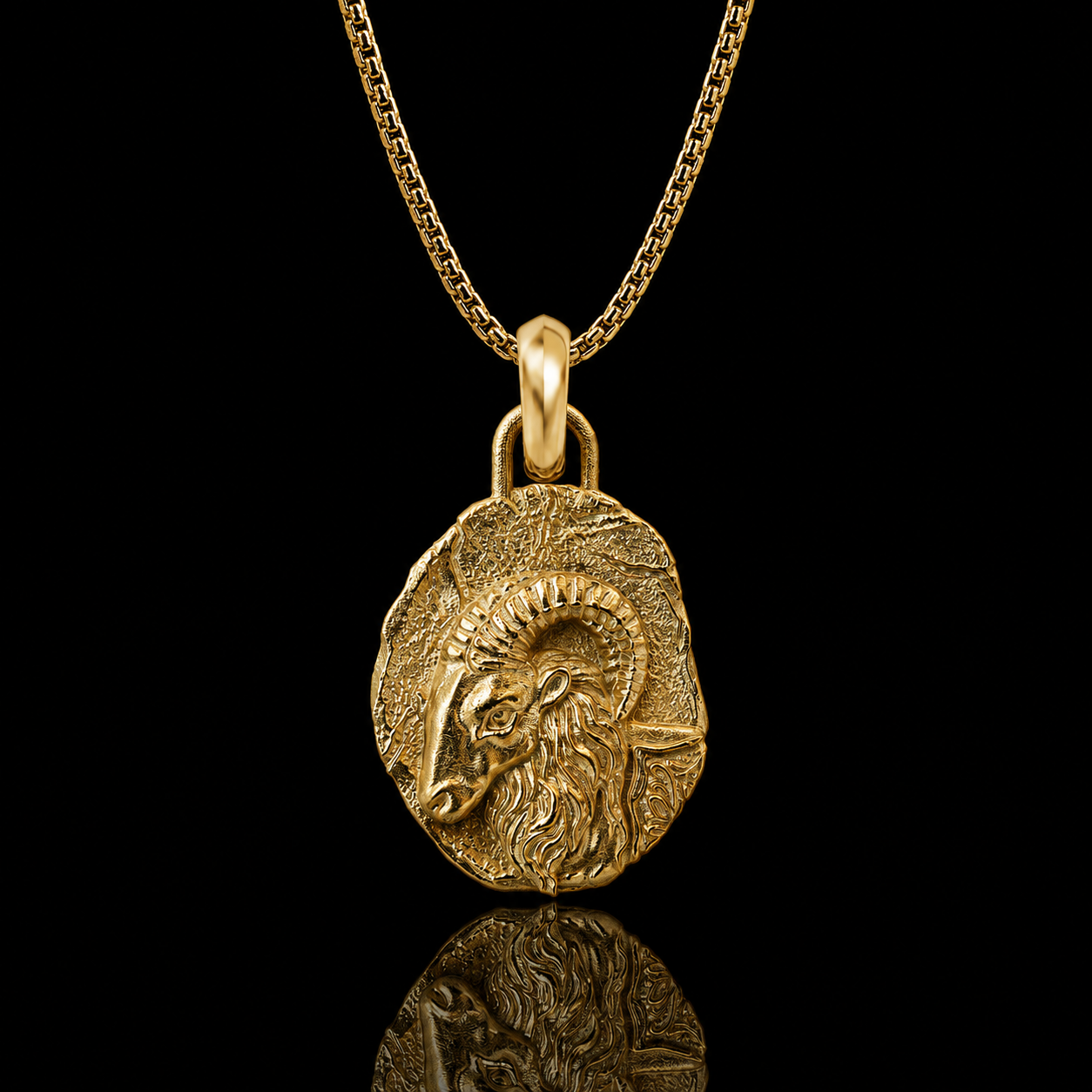 A gold pendant necklace featuring a textured ram's head design, suspended from a gold chain against a solid black background with a soft reflection below.