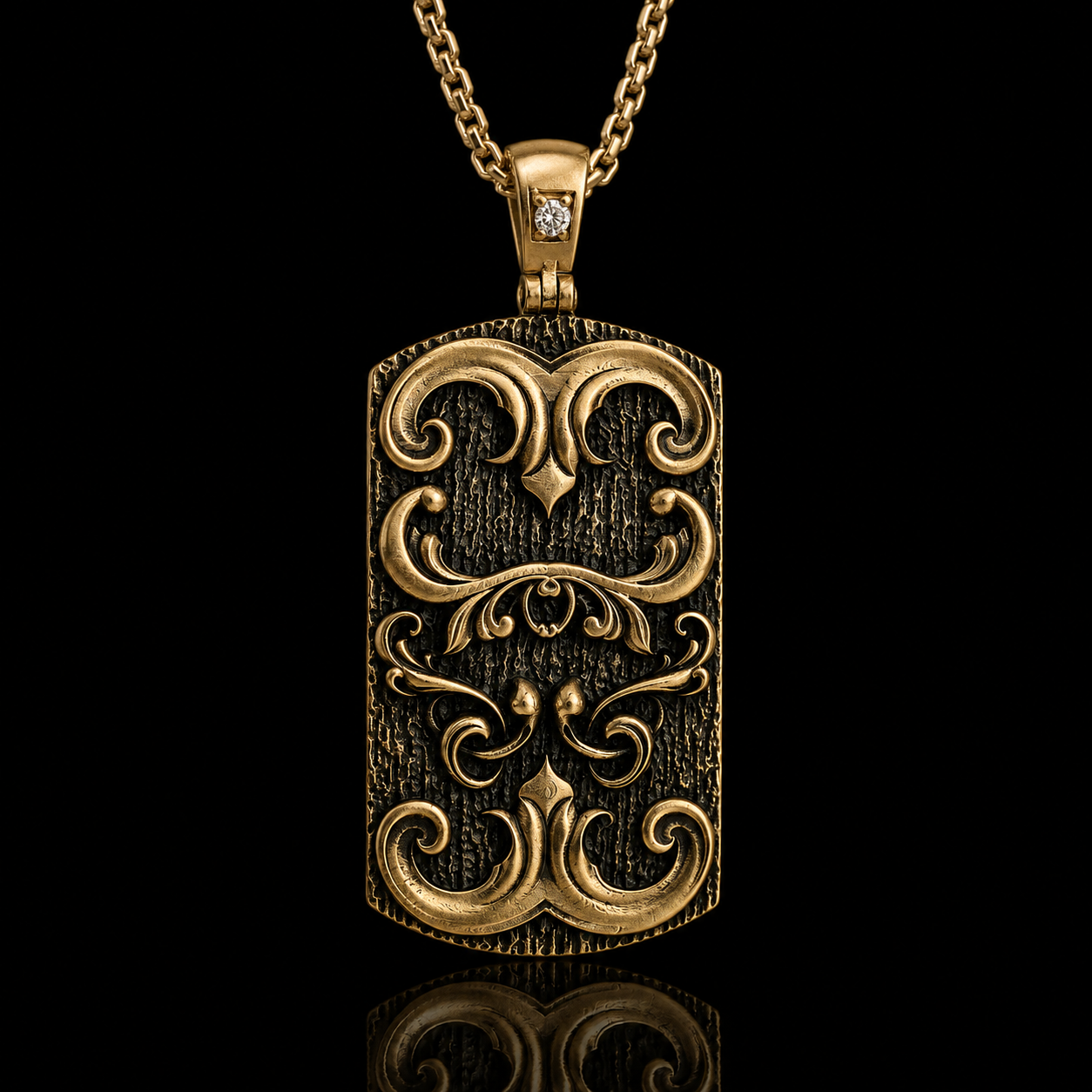 An ornate gold-toned rectangular pendant necklace with a textured finish, embossed scrollwork, and a small clear stone set in the top, shown on a black background with a reflection below.