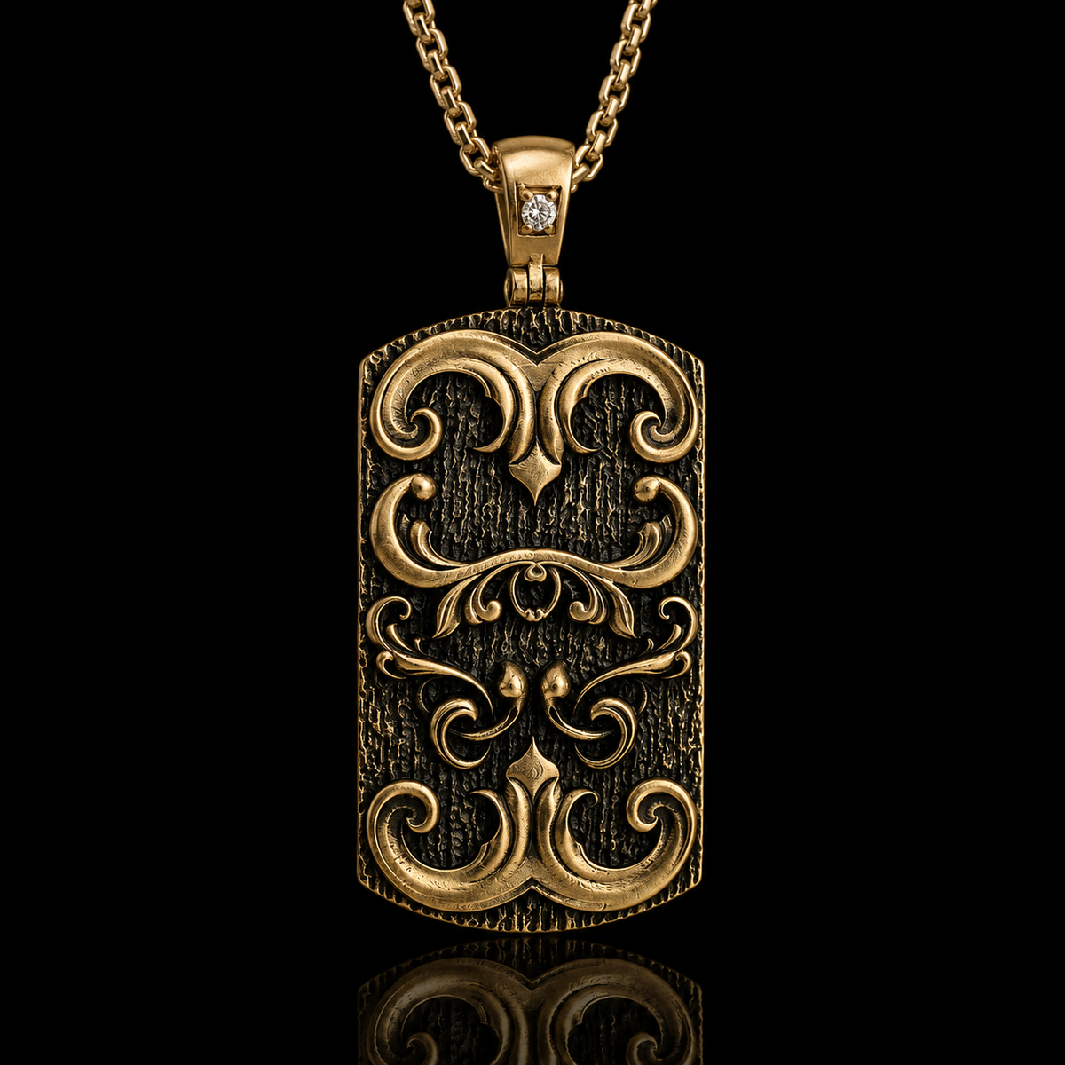 An ornate gold-toned rectangular pendant necklace with a textured finish, embossed scrollwork, and a small clear stone set in the top, shown on a black background with a reflection below.