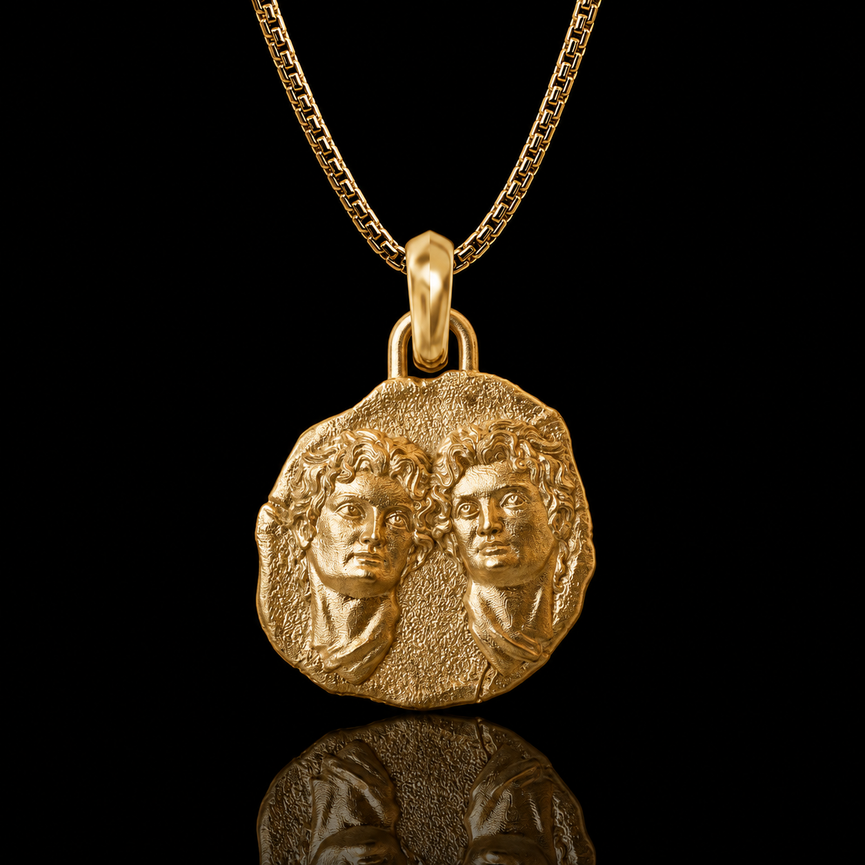 A gold pendant necklace featuring an ancient coin-style medallion with two embossed classical busts, suspended from a gold chain against a solid black background with a reflection below.