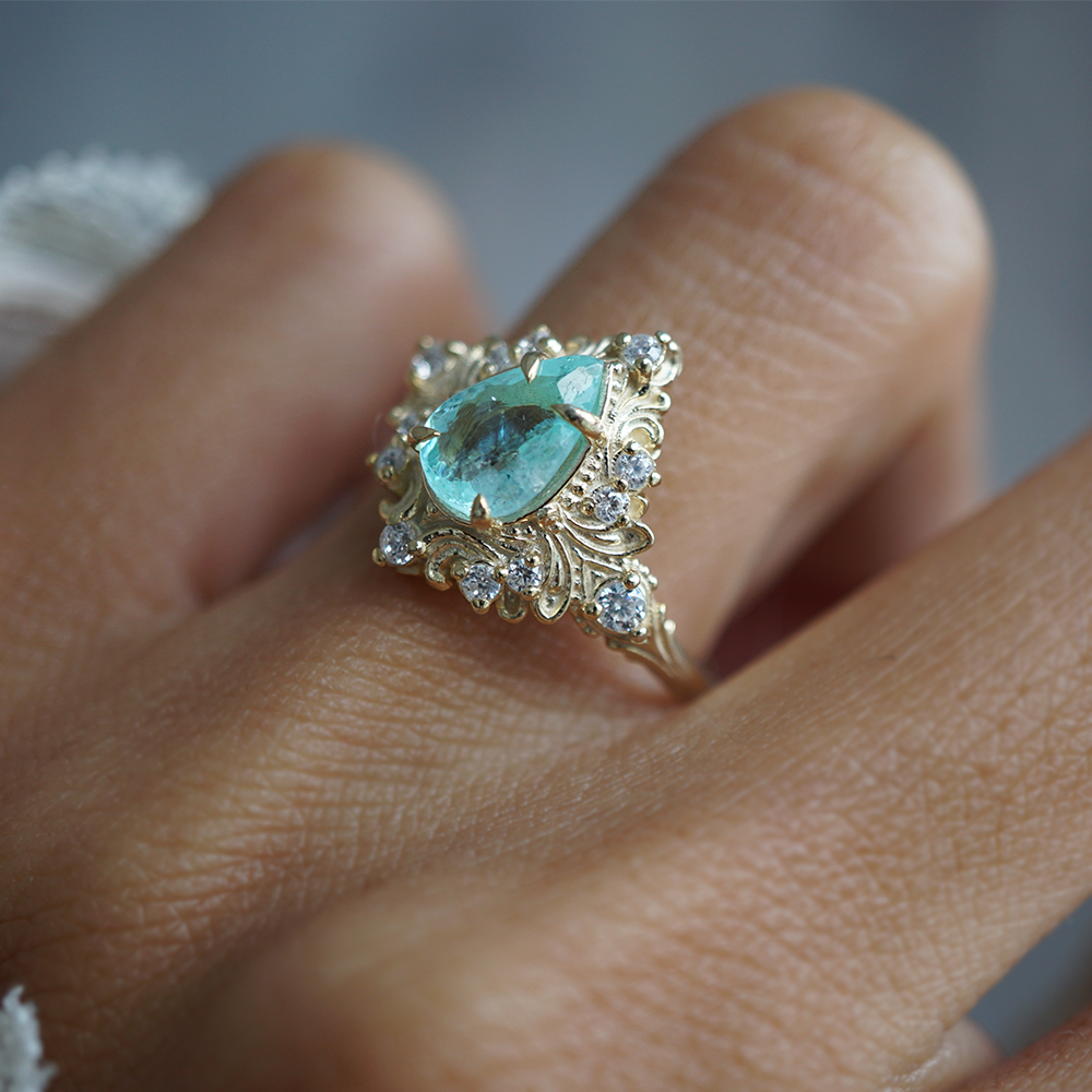 A close-up of a gold ring with a pear-cut turquoise paraiba tourmaline center stone and small diamonds, worn on a finger.