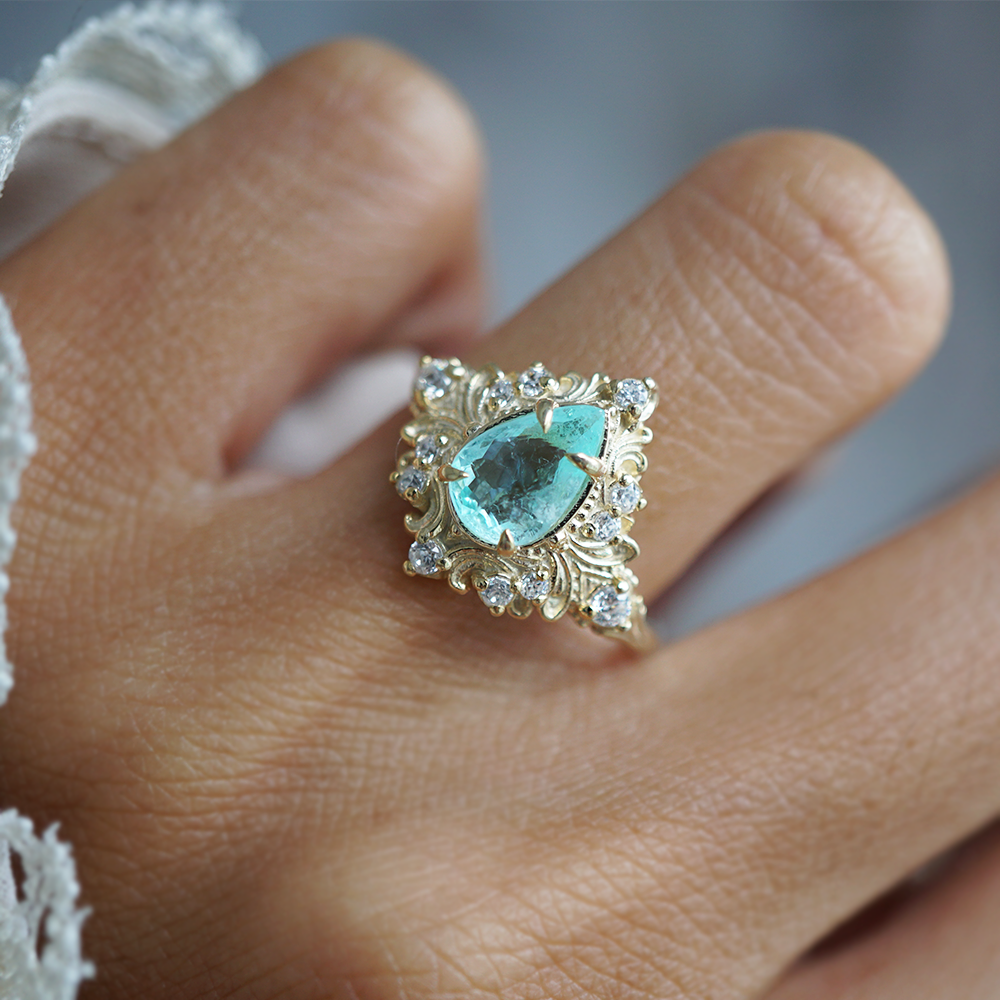 A close-up of a hand wearing a gold ring with a central turquoise-colored paraiba tourmaline pear gemstone surrounded by small diamonds.