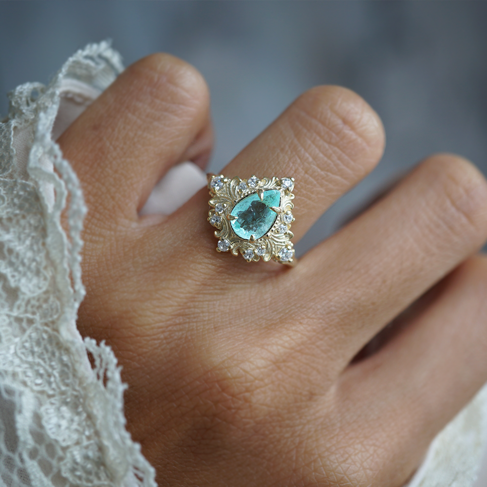 A close-up of a hand wearing a vintage-style gold ring with a pear-cut paraiba tourmaline center stone and small clear accent stones.