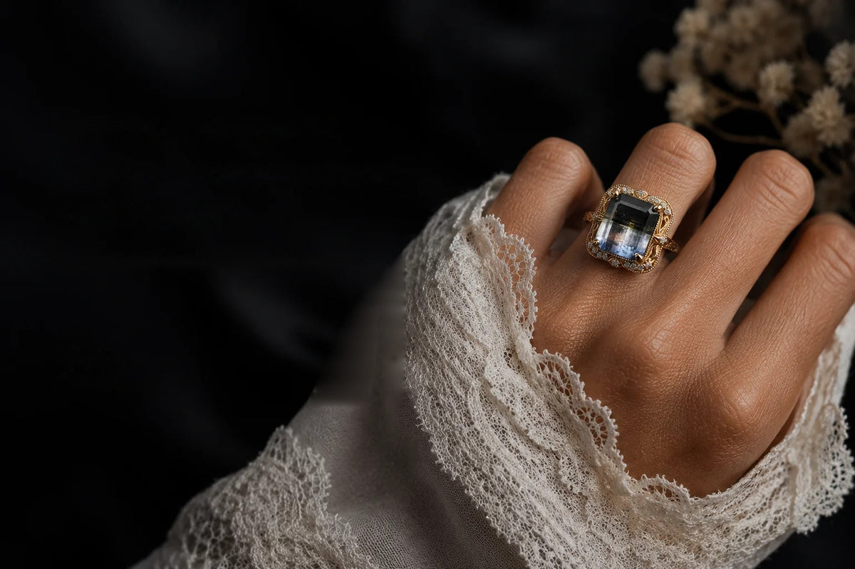 A hand in a lace-cuffed sleeve wearing a Tippy Taste gemstone ring, against dark fabric