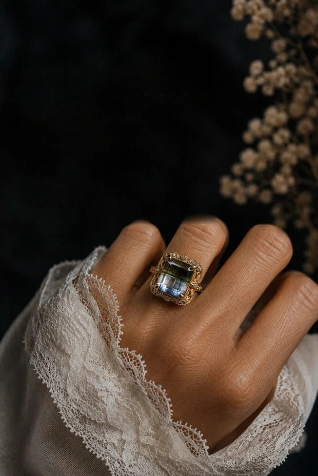 A hand in a lace-cuffed sleeve wearing a Tippy Taste gemstone ring, against dark fabric