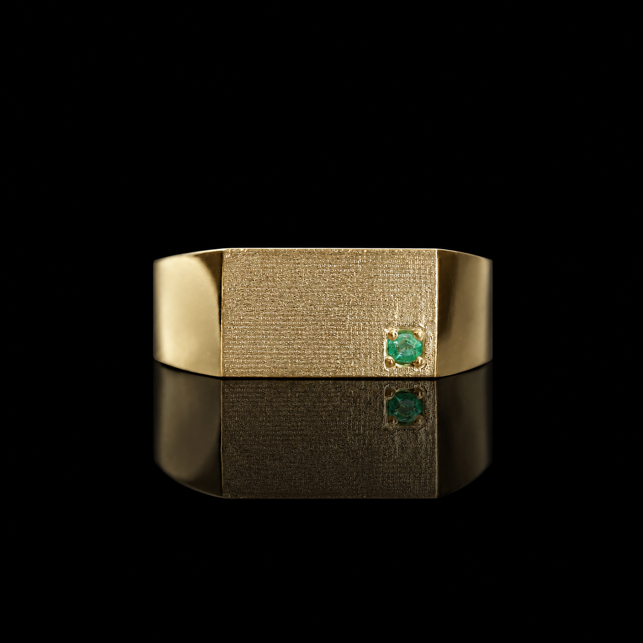 A gold signet-style ring with a textured rectangular face and a small round paraiba tourmaline gemstone on the right side, photographed on a black reflective surface.