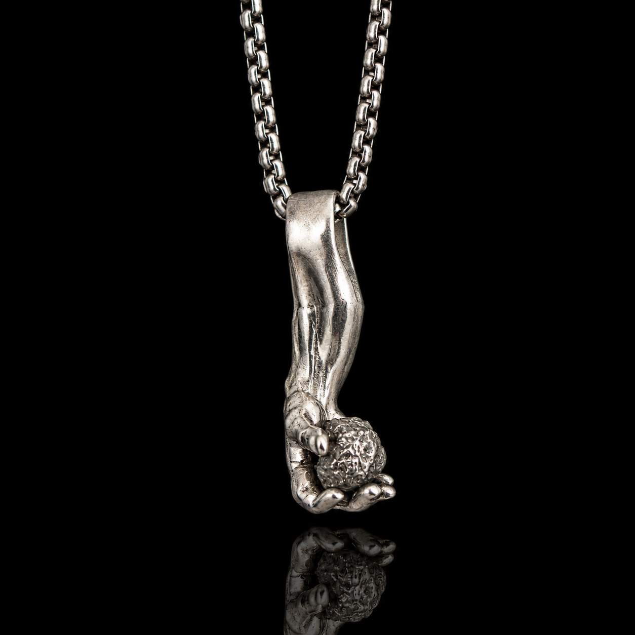 A silver pendant necklace featuring a sculpted hand holding a natural raw diamond, suspended from a silver box chain against a solid black background with a soft reflection below.