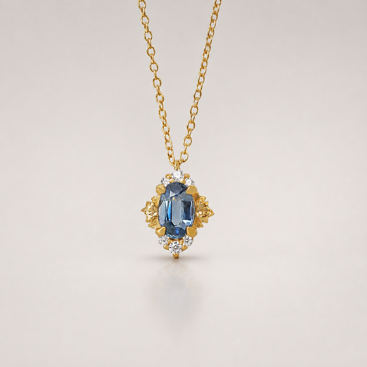 A gold pendant necklace featuring an oval-cut blue sapphire surrounded by small diamonds and gold floral details, suspended from a delicate gold chain against a light gray background.